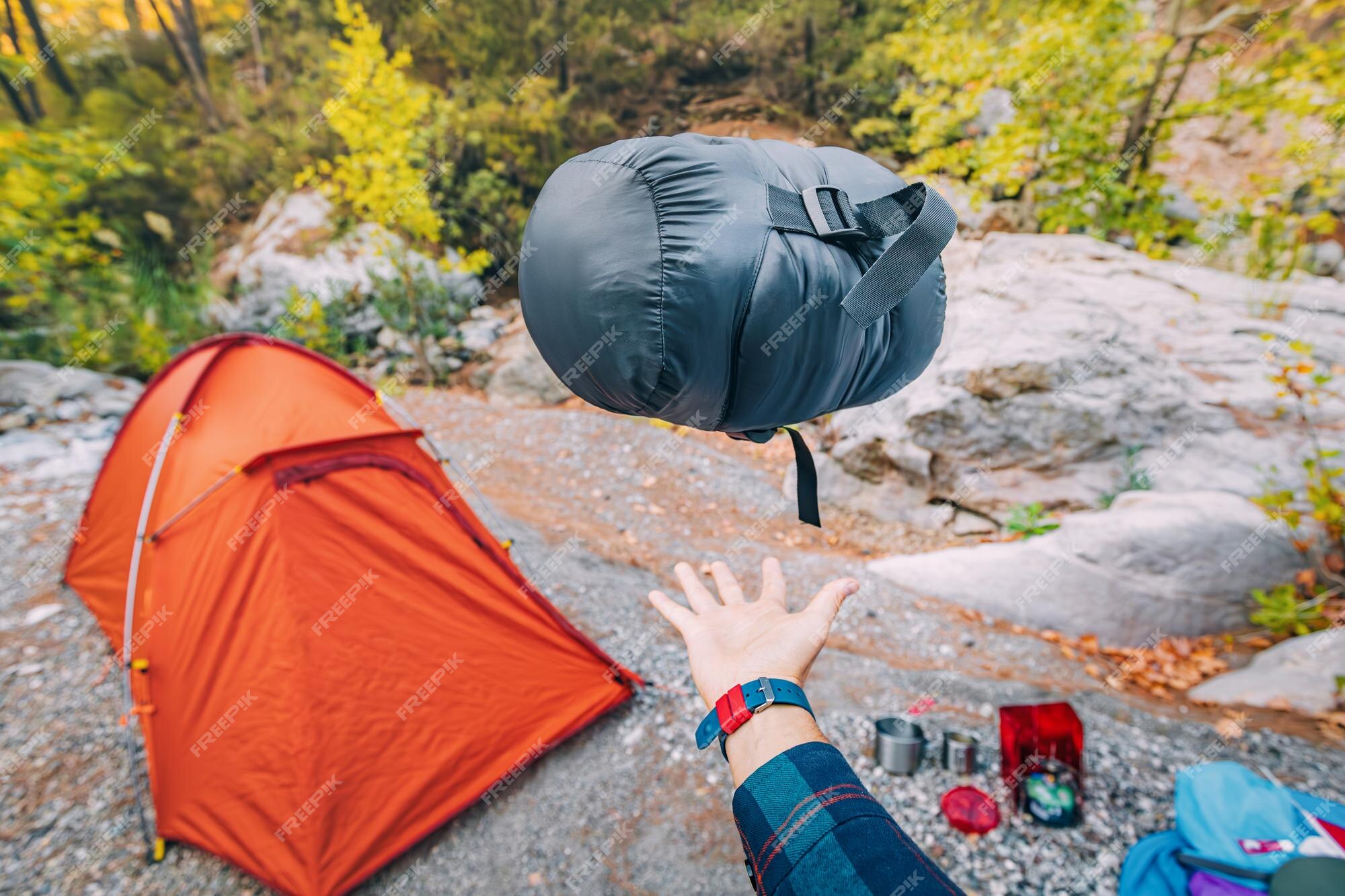 Collapsible camping tent, sleeping bags, and cooking gear arranged around a campfire in a forest setting.